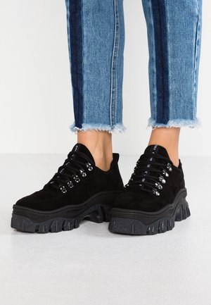 Schwarze Sneakers mit dicken Sohlen und silbernen Ösen, getragen zu blauen, gekürzten Jeans mit dunklen Seitenstreifen und ausgefransten Säumen.