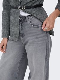 Primer plano de una persona que lleva vaqueros de pierna ancha grises, blazer gris texturizado ceñido con cinturón negro fino y anillo plateado en la mano derecha.