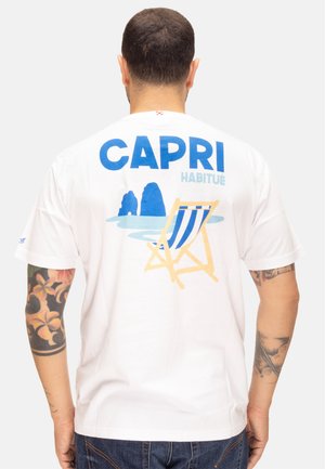 Uomo con tatuaggi che indossa una t-shirt bianca con la scritta blu "CAPRI", il testo azzurro "HABITUÉ" e una grafica di una sedia da spiaggia sul retro.