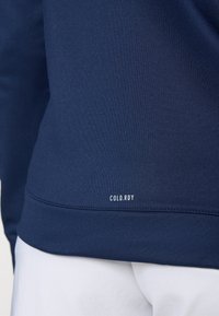 Close-up of a person wearing a navy blue sweatshirt with "COLD.RDY" text near the hem and white pants.