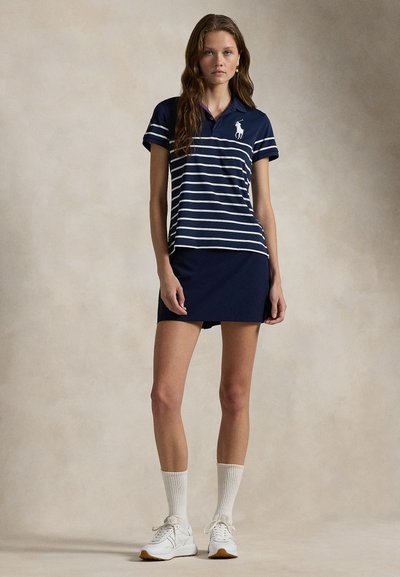 Giovane donna che indossa una polo a righe blu navy con un logo bianco, gonna blu navy, calzini bianchi e sneakers bianche, in piedi su uno sfondo neutro.
