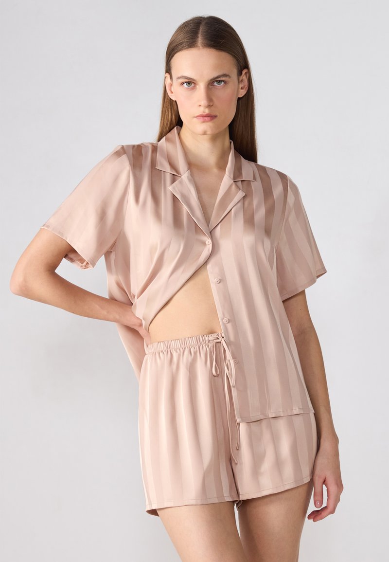 Ljusrosa satinpyjamas med korta ärmar och lös passform. Har vertikala ränder, en krage och shorts med elastisk dragsko.
