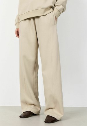 Bēša platām kājām bikses kopā ar brūnām smailām kurpēm un atbilstošu bēšu sporta krekli, redzamas no vidukļa līdz kājām.