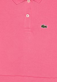 Polo rose en coton texturé avec un plastron à deux boutons et un petit logo de crocodile vert sur la poitrine gauche.
