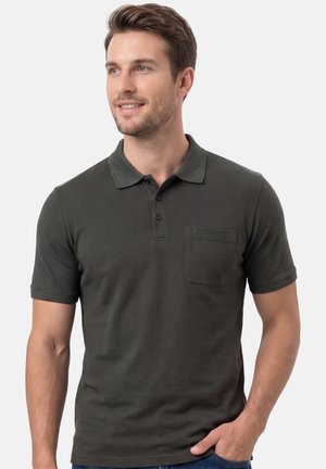 Mężczyzna w ciemnoszarej koszulce polo z krótkim rękawem i przednią kieszenią, patrzący lekko na bok, ręka w kieszeni jeansów, neutralne tło.