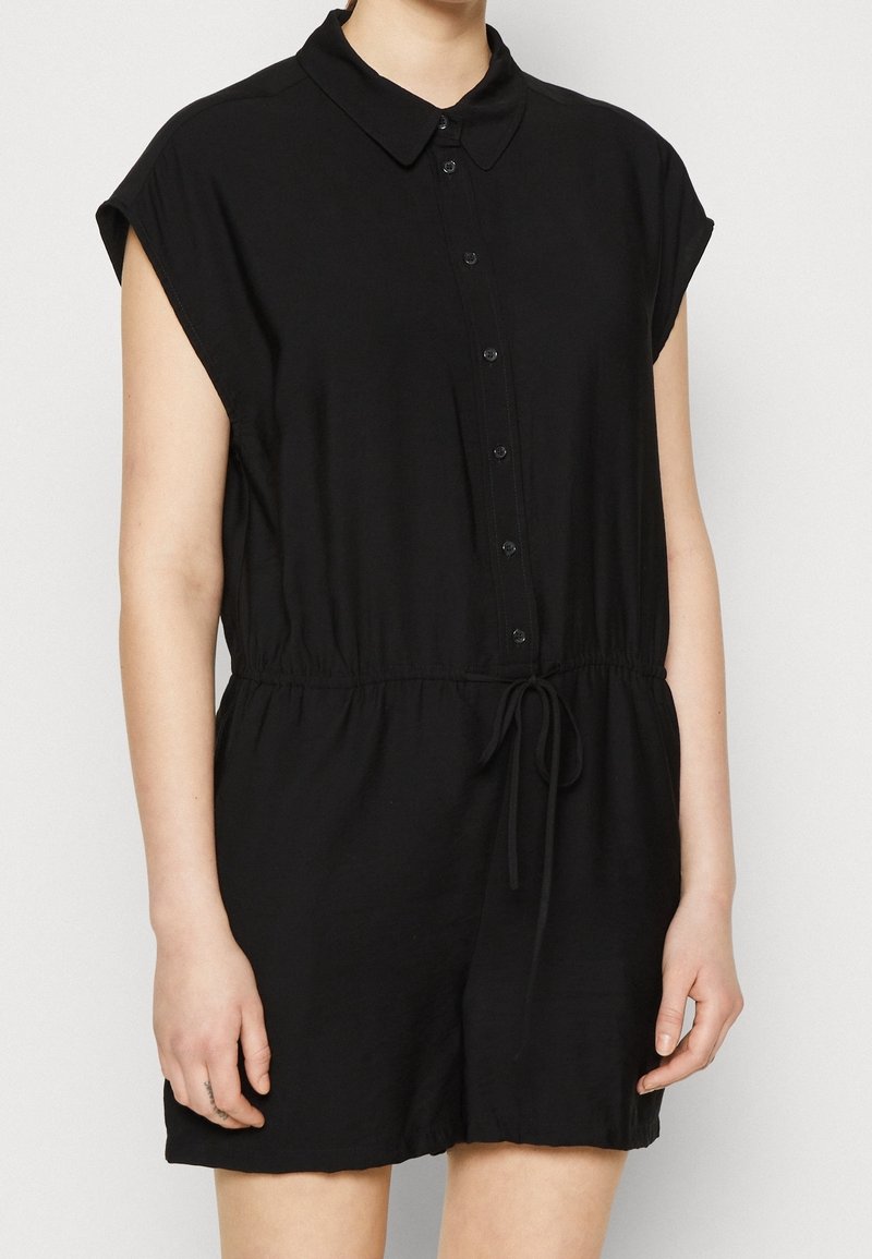 Femme portant une combinaison noire à manches courtes, boutonnée, avec un col et un cordon à la taille, debout devant un fond clair uni.