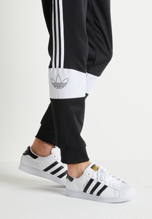 Svarta joggers med vita detaljer och tre vita ränder på sidorna, kombinerade med vita sneakers med svarta ränder och en guldfärgad logotyp.