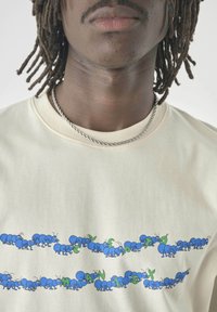 Close-up of person's neck and chest wearing a beige shirt with blue ants carrying green letters forming "PANGAIA X" on front.