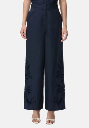 Donna che indossa pantaloni a gamba larga blu navy con ricamo floreale traforato vicino all'orlo, abbinati a tacchi bianchi open-toe.