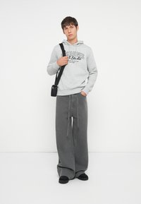 Sudadera gris con logo, combinada con pantalones de mezclilla gris de pierna ancha. Un bolso y zapatos negros completan el conjunto. Diseño sencillo y casual.