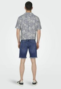 Camisa de flores de manga corta, de color gris claro, con patrones de palmeras y veleros, combinada con pantalones cortos de mezclilla. Se ven los puños enrollados y los bolsillos traseros.