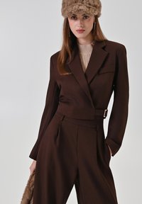 Bruine op maat gemaakte blazer met een cropped ontwerp en een ceintuurdetail, gecombineerd met hooggetailleerde bruine broek. Textured beige trui eronder.