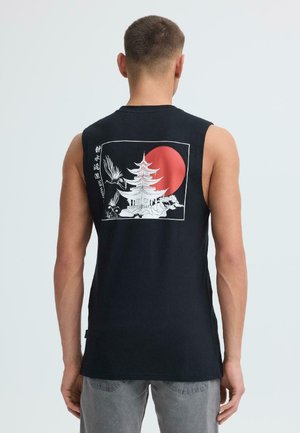 Homme portant un débardeur noir avec un motif de pagode blanche, d'oiseau et de soleil rouge dans le dos, debout devant un fond clair uni.