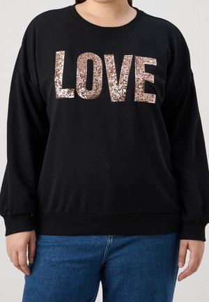 Persoon die een zwarte longsleeve draagt met grote rosegouden pailletten "LOVE" tekst, gecombineerd met blauwe jeans, tegen een effen achtergrond.