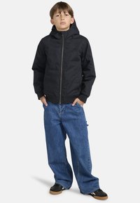 Sort hættetrøje med lynlås foran, parret med løstsiddende blå jeans og sorte sneakers. Enkel design uden synlige mønstre.