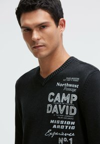 Schwarzer Pullover mit V-Ausschnitt und geripptem Kragen. Mit weißem und grauem Drucktext, darunter "CAMP DAVID" und "Northwest Passage".
