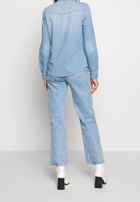 Chemise en denim bleu clair avec un col boutons, manches longues et détail de la poche au dos, assortie à un jean droit bleu clair et des bottes blanches à talons.