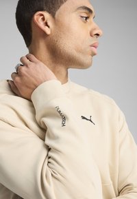 Sudadera beige con un logo negro de Puma en el pecho y el texto "PUMATECH" en la manga. Tejido suave con un ajuste relajado.