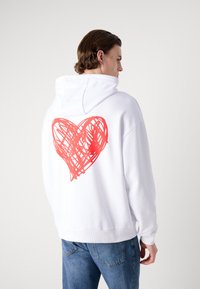 Felpa bianca con cappuccio, caratterizzata da una grande grafica a forma di cuore rosso disegnata a mano sul retro. Tessuto morbido, vestibilità comoda, polsini a costine.