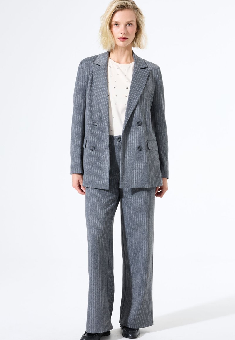 Conjunto de blazer y pantalones a rayas gris, con corte entallado, blazer de doble botonadura, pantalones de frente plano, combinado con una blusa crema con pequeñas embellishments.