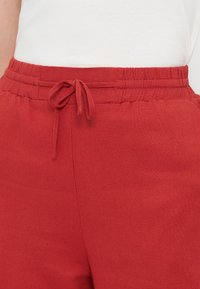 Κοντινό πλάνο κόκκινου παντελονιού με ελαστική μέση και δεμένη υφασμάτινη κορδονίτσα, φορεμένο με λευκό τοπ που είναι μέσα στην μέση.