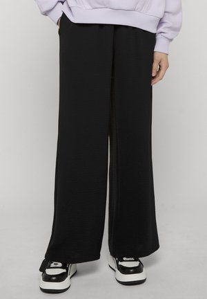 Person wearing loose black pants, black and white sneakers, and a light purple sweatshirt with hands in pockets.