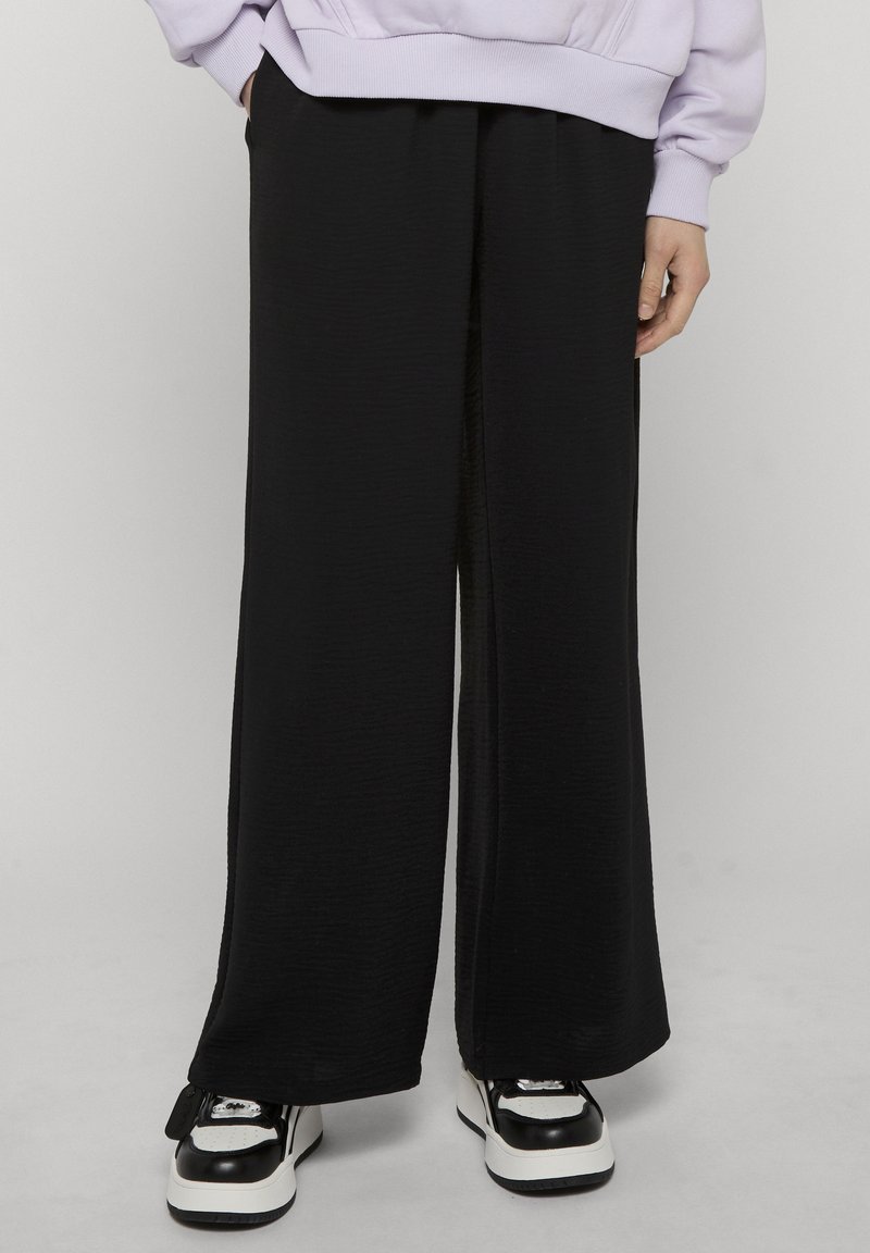 Personne portant un pantalon noir ample, des baskets noires et blanches, et un sweat-shirt violet clair les mains dans les poches.