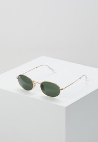 Occhiali da sole in metallo dorato con lenti ovali verdi, aste metalliche sottili e punte trasparenti. Le montature presentano un design minimalista e arrotondato.