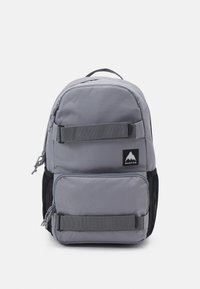 Gray fabric backpack with a rounded top, front zip pocket, mesh side pockets, and vertical straps. Features a logo patch in black and white.