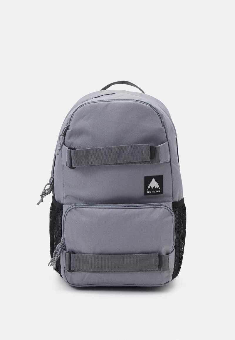 Gray fabric backpack with a rounded top, front zip pocket, mesh side pockets, and vertical straps. Features a logo patch in black and white.