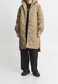 Quiltad khaki lång kappa med huva, utrustad med tryckknappar och en avslappnad passform. Kombinerad med en svart t-shirt och vida byxor.