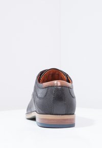 Chaussure en cuir noir texturé avec une forme arrondie, présentant un accent en cuir marron au talon et une semelle en bois.