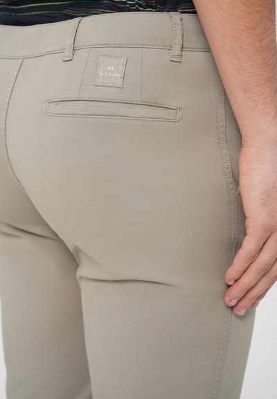 Beżowe spodnie o gładkiej fakturze, z tylną kieszenią oraz metką z napisem „PS Paul Smith”. Krój jest dopasowany i elegancki.