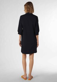 Svart klänning med trekvartsärmar, rak skärning och mjuk textur. Har en liten krage och är knälång, bärs med sandaler.