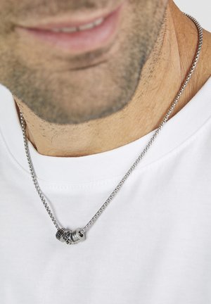 Silver metal necklace with interlinked circular and textured pendants, resting on a white t-shirt. The chain is woven and smooth.