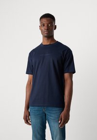 Līklā zila kokvilnas T-kreklu ar apaļu izgriezumu un īsām piedurknēm, ar neuzkrītošu "Scotch & Soda" logo uz krūtīm. Valkāts ar zilām džinsām.