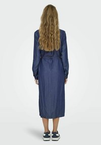 Denim midi haljina s dugim rukavima, gumbima na zapešćima, strukirana i sa ravnim rubom, u paru s tamnim tenisicama.