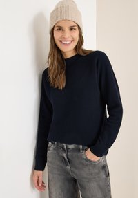 Maglione lavorato a maglia blu navy con collo rotondo, texture a coste e maniche lunghe, abbinato a jeans grigio chiaro. Berretto beige lavorato a maglia.