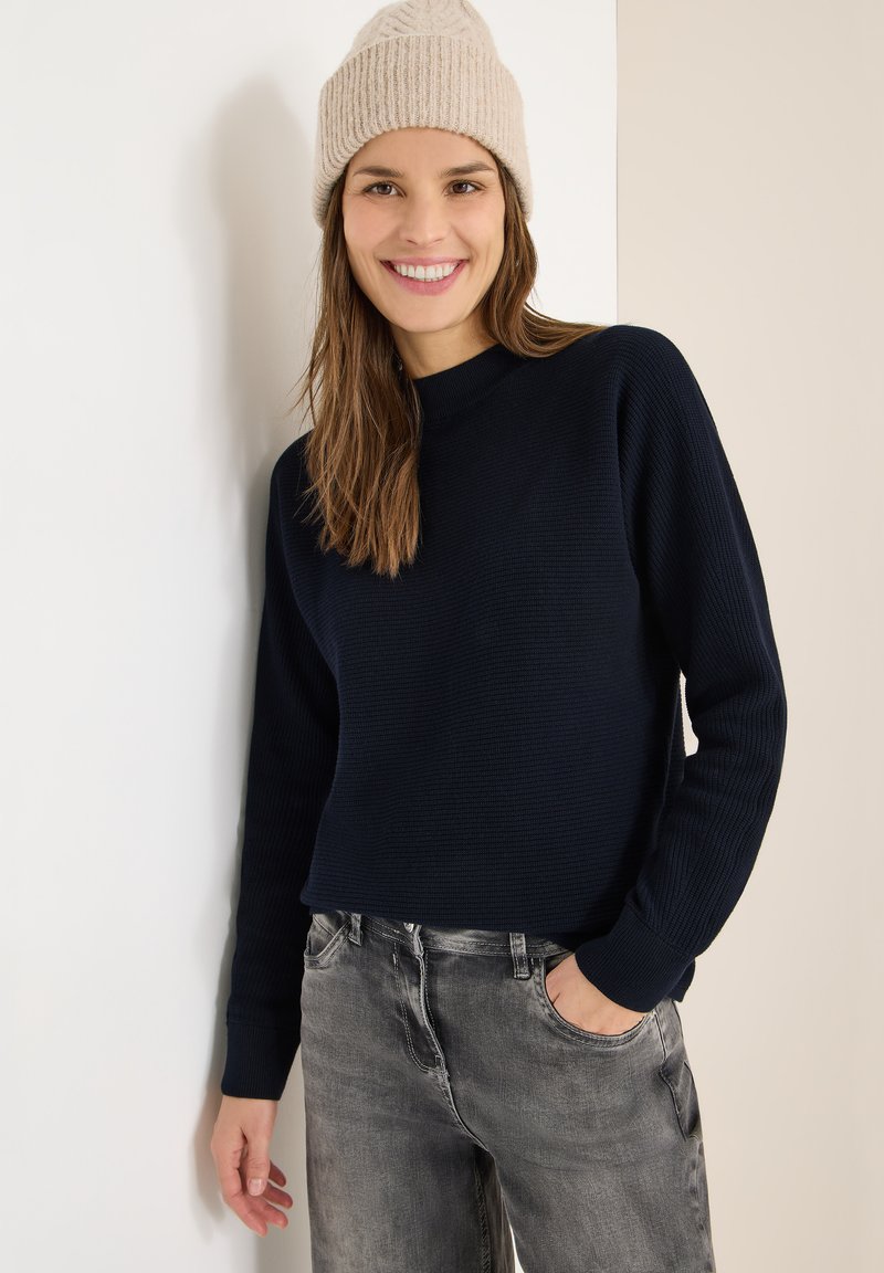 Maglione lavorato a maglia blu navy con collo rotondo, texture a coste e maniche lunghe, abbinato a jeans grigio chiaro. Berretto beige lavorato a maglia.