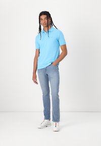 Camisa polo azul clara com mangas curtas, apresentando um colarinho e um logótipo bordado, combinada com uns jeans de ganga claros e sapatilhas brancas.