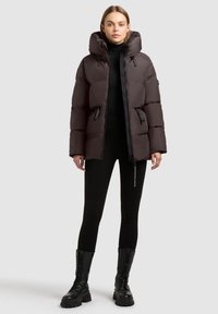 Brun puffjakke med hætte, der har en lynlåslukning foran, to sidelommer og sorte detaljer. Båret over en sort top og leggings.