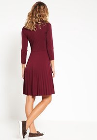 Burgundymidi-klänning med en figurnära överdel, trekvartsärmar och en plisserad kjol. Tillverkad av mjukt tyg, med subtil textur.