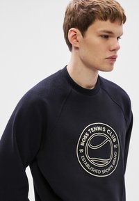 Tumši zila sporta džempera ar ribotu apaļu apkakli, struktūrētu audumu un lielu izšūtu tenisa kluba logo uz krūtīm.