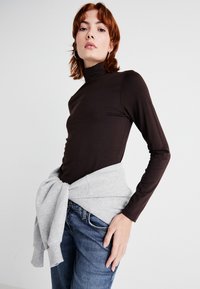 Ruda ilga rankovė turtlenecko tipo palaidinė, pagaminta iš minkštos medžiagos, derinama su pilku džemperiu, surištu aplink liemenį. Mėlyni džinsai užbaigia įvaizdį.