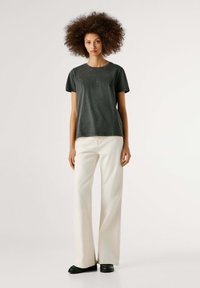 Tumeda halli lühikeste varrukatega T-särk, millel on reljeefne tekst, kombineeritud laia jala kreemikate pükstega ja mustade kingadega, ühtlase tausta ees.