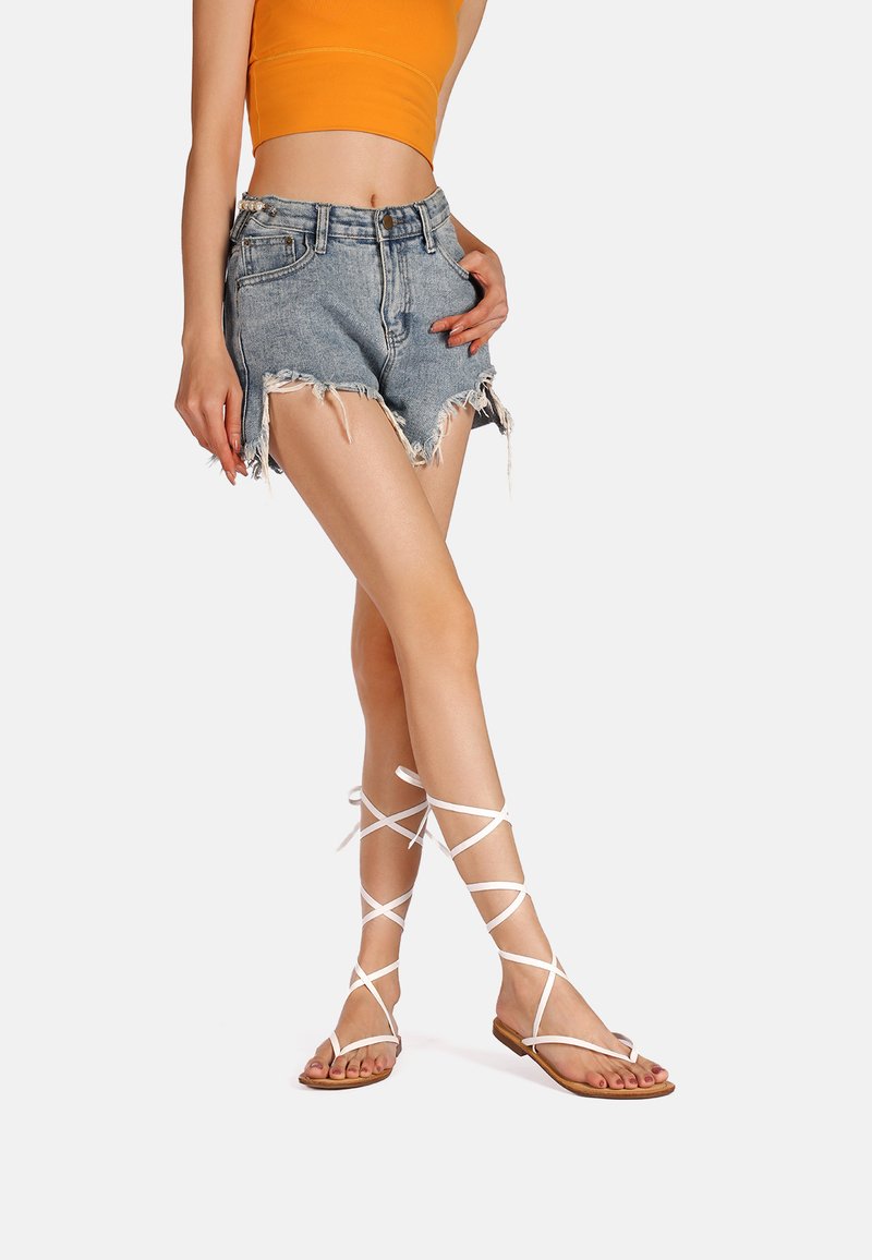 Sandali bianchi con lacci e suola piatta, caratterizzati da sottili cinturini che avvolgono le gambe. Abbinati a pantaloni corti in denim sfilacciati e un top corto arancione.