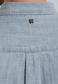 Camisa de color azul claro con una tela texturizada, que presenta una espalda plisada y una pequeña etiqueta con logo negro y dorado en el cuello.