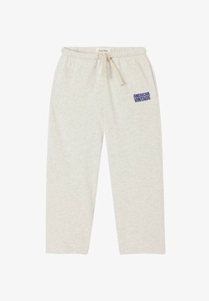 Persona giovane che indossa una t-shirt blu "American Vintage", pantaloni beige chiaro, scarpe beige slip-on e una sciarpa annodata intorno al collo, in piedi in modo rilassato.