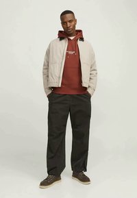 Giacca bomber beige chiaro sopra un felpa bordeaux, abbinata a pantaloni neri larghi e scarpe marroni con suola in gomma.