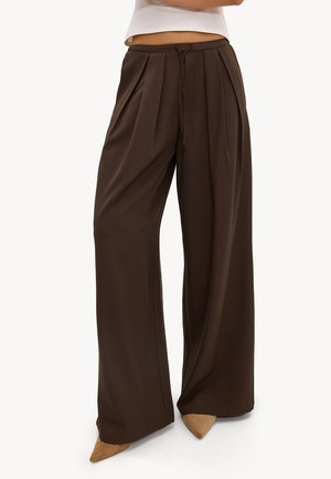 Personne portant un pantalon marron foncé taille haute à jambes larges avec des plis et un cordon, associé à des chaussures beiges pointues et un haut blanc.